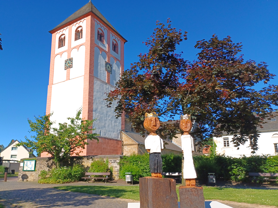 Foto von zwei geschnitzte Holzfiguren, die König und Königin darstellen - Kirche St. Pankratius im Hintergrund
